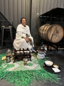 A woman wearing Ethiopian clothing sitting in front of small cups and an Ethiopian coffee pot and heater